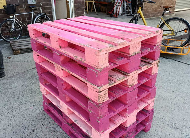 Gestapelte, pink lackierte Holzpaletten auf einem Betonboden vor einer Backsteinwand mit Fahrrädern im Hintergrund