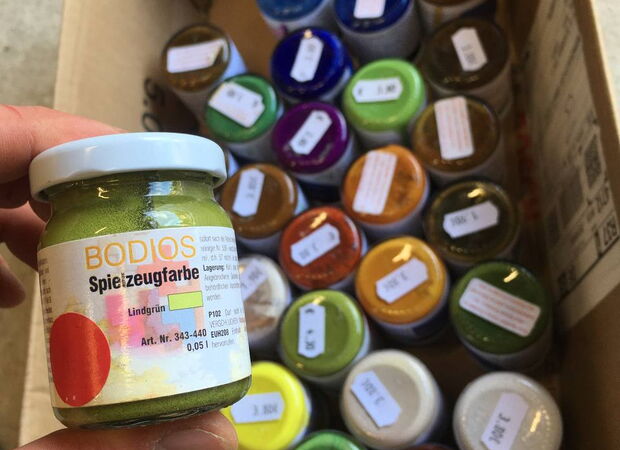 Nahaufnahme eines Glases mit grüner Spielzeugfarbe der Marke BODIOS vor mehreren weiteren Farbdosen in einer Kiste