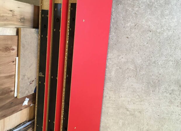 Mehrere übereinanderliegende Hängeregale mit roter Oberfläche liegen auf einem Betonboden neben einer Holzplatte.