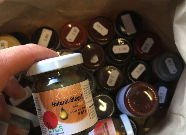 Hand hält kleines Glas mit Aufschrift "Naturöl-Siegel" vor mehreren Gläsern mit weißen Etiketten in einer Kiste