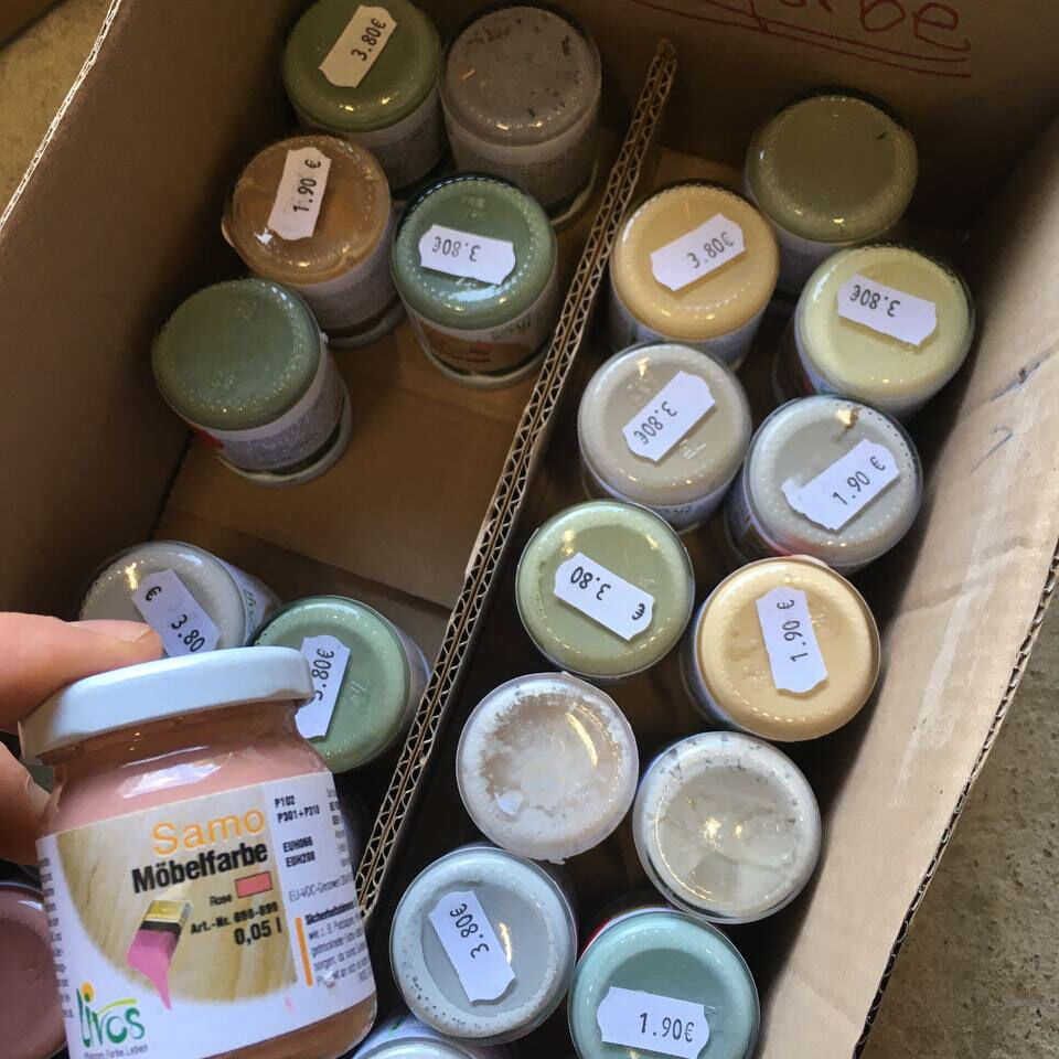 Mehrere kleine Farbdosen mit Preisschildern in einem Karton, eine Dose mit der Aufschrift 'Same Möbelfarbe' wird von einer Hand gehalten.