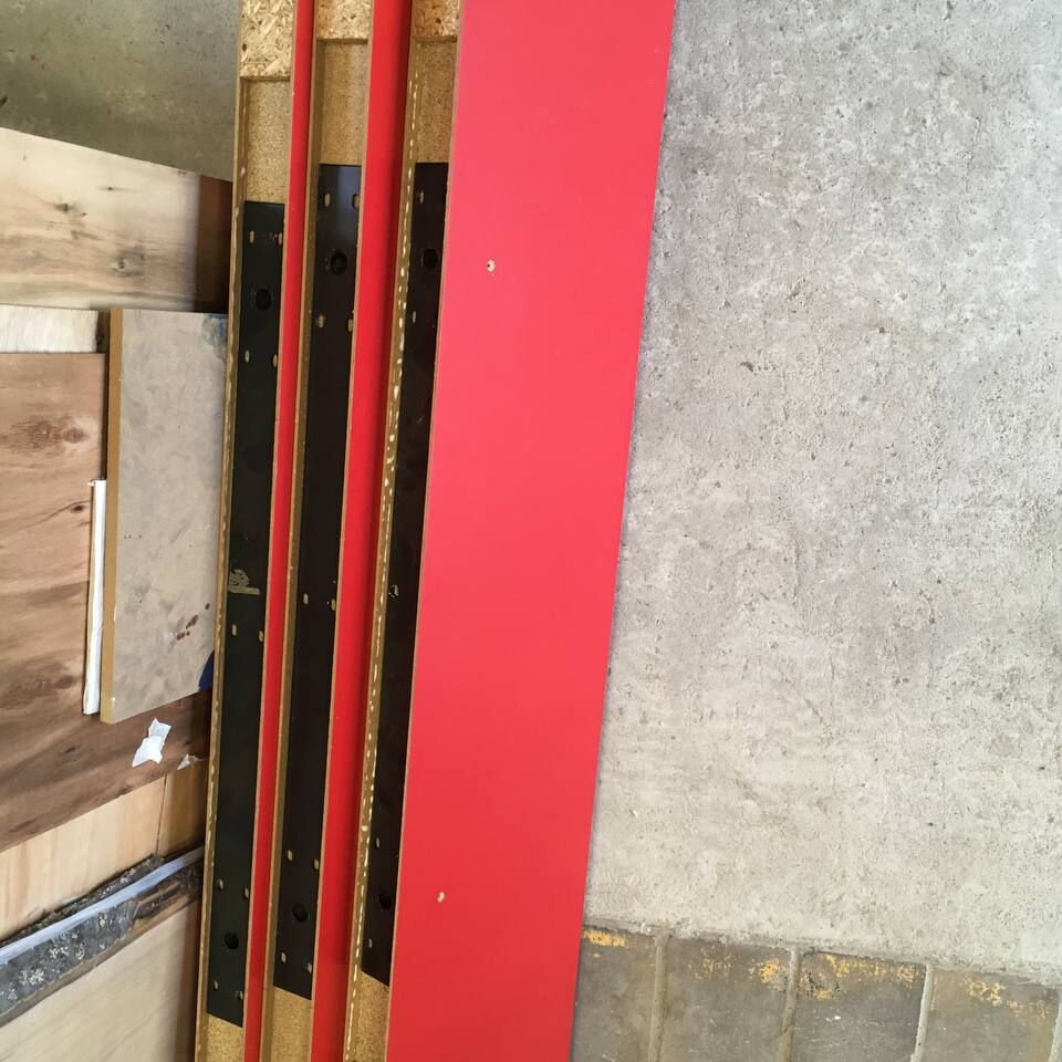 Mehrere übereinanderliegende Hängeregale mit roter Oberfläche liegen auf einem Betonboden neben einer Holzplatte.